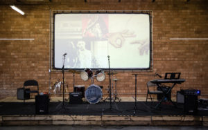 empty band stage featuring mic stands, a drum set, keyboards, amps, and a wall projection screen